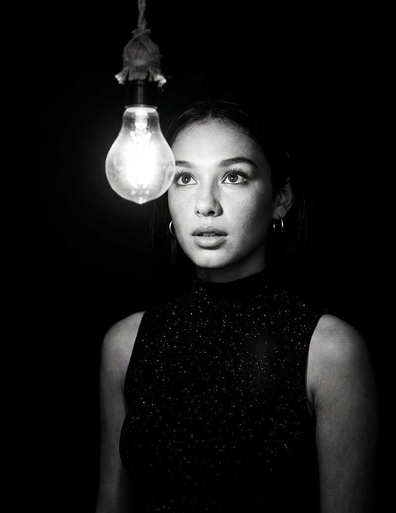 Nova Rai beside a single bare light bulb in the dark, looking past it, in black and white