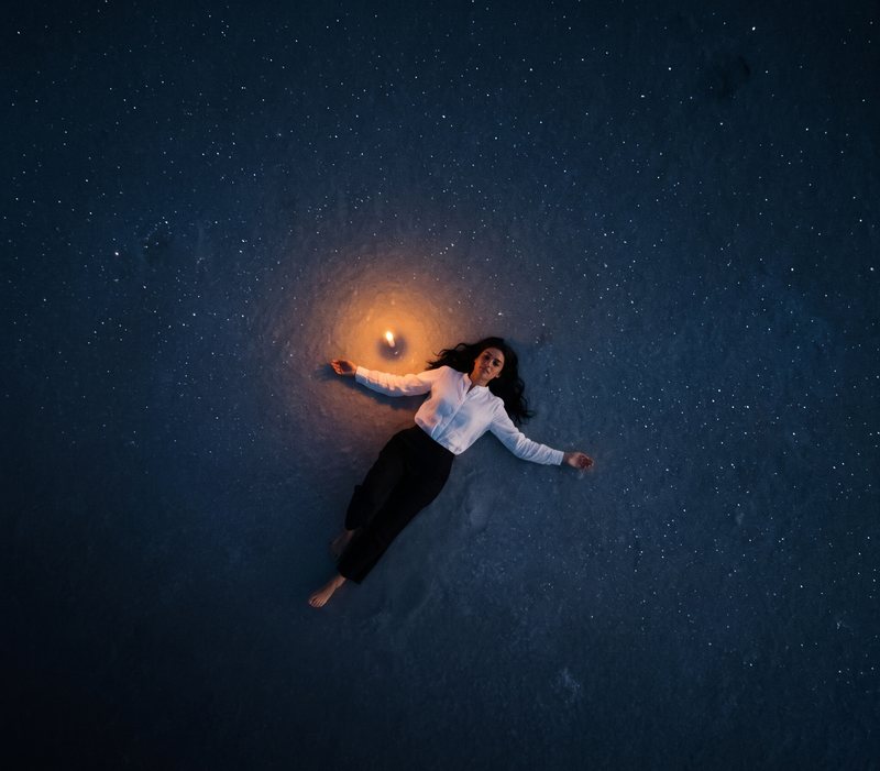 Nova Rai lying on a dark floor full of reflected stars, a small candle beside her