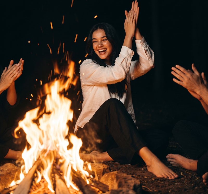Nova Rai laughing and clapping by a bonfire at night, other hands clapping around her