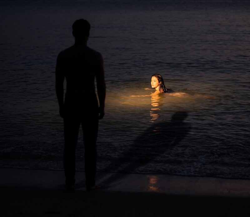 Nova Rai swimming far out in a dark sea at night, a man's shadow waiting at the waterline