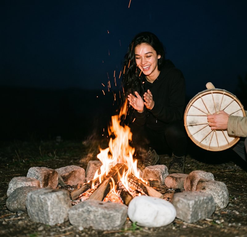 Nova Rai laughing by a fire ring of stones, a hand drum beside her